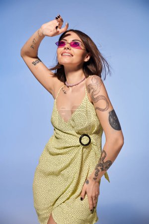 A brunette woman in a stunning yellow dress proudly shows off her intricate arm tattoos in a studio setting.