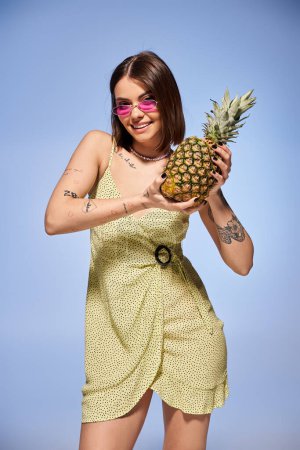 A brunette woman in a yellow dress joyfully holds a pineapple in a studio setting.
