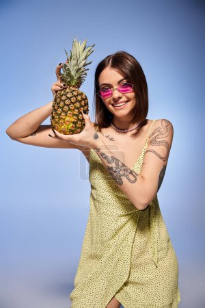 A young woman with brunette hair poses in a studio setting wearing a bright yellow dress, holding a vibrant pineapple.