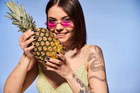 Eine stylische junge Frau mit brünetten Haaren hält eine Ananas in der Hand, während sie im Studio eine trendige Sonnenbrille trägt.