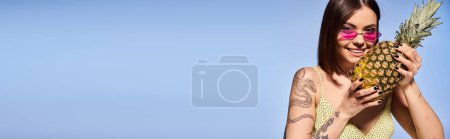 Téléchargez les photos : Une jeune femme aux cheveux bruns tenant un ananas tout en portant des lunettes de soleil roses dans un cadre studio vibrant et joyeux. - en image libre de droit