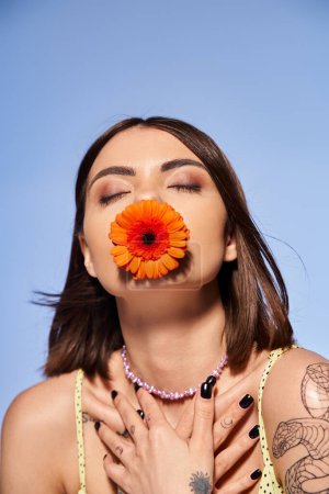 A young woman with brunette hair elegantly holds a delicate flower in her mouth in a studio setting.