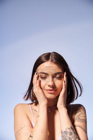 Téléchargez les photos : Une jeune femme avec des tatouages et des cheveux bruns, tenant ses mains sur son visage dans un cadre de studio. - en image libre de droit