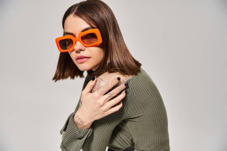 Une jeune femme brune pose dans un pull vert vif et des lunettes de soleil orange dans un cadre studio.