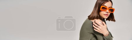 Téléchargez les photos : Une jeune femme aux cheveux bruns pose en toute confiance dans un studio, portant un pull vert vif et des lunettes de soleil orange à la mode. - en image libre de droit