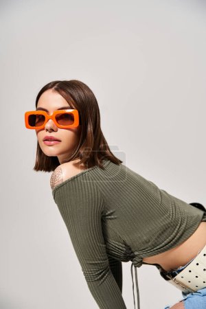 Téléchargez les photos : Une femme brune élégante debout en toute confiance dans un haut vert et des lunettes de soleil orange tendance. - en image libre de droit