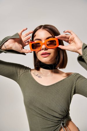 A stylish young woman with brunette hair wearing a green shirt and orange sunglasses in a studio setting.