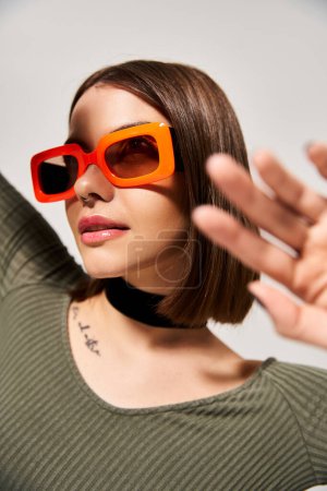 A stylish young woman with brunette hair wearing orange sunglasses and a green shirt in a studio setting.