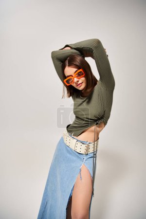 A stylish young woman with brunette hair poses confidently in a skirt and sunglasses in a professional studio setting.