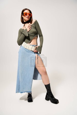 A stylish brunette woman strikes a pose in a skirt and boots in a studio setting.
