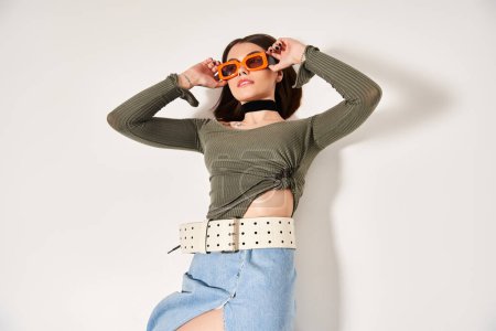 A young woman with brunette hair wears a green shirt and stylish orange sunglasses in a studio setting.