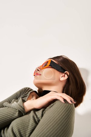 A young woman with brunette hair poses stylishly in a studio, wearing a vibrant green sweater and trendy orange sunglasses.