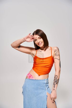 Une jeune femme aux cheveux bruns pose en toute confiance dans un studio portant un haut orange et une jupe bleue fluide.