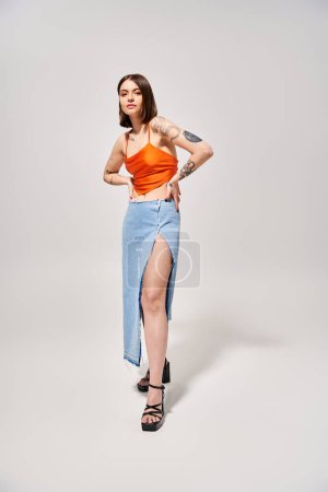 A stylish young woman with brunette hair stands gracefully in a studio wearing an orange top and a flowing blue skirt.