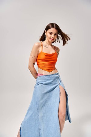 A young woman with brunette hair poses gracefully in a studio, wearing an orange top and a flowing blue skirt.