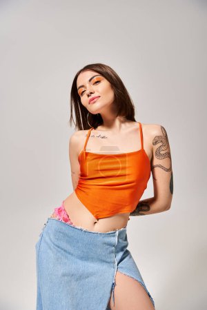 A stylish young woman with brunette hair poses in a studio wearing an orange top and a flowing blue skirt.