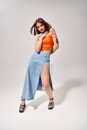 Photo for A young woman with brunette hair poses in a studio wearing an orange top and blue skirt, exuding elegance and grace. - Royalty Free Image