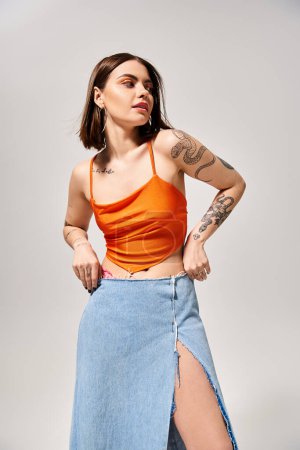 A stylish young woman with brunette hair wearing an orange top and blue skirt in a studio setting.