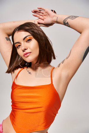 A young woman with brunette hair showcases intricate tattoos on her arms and chest in a studio setting.