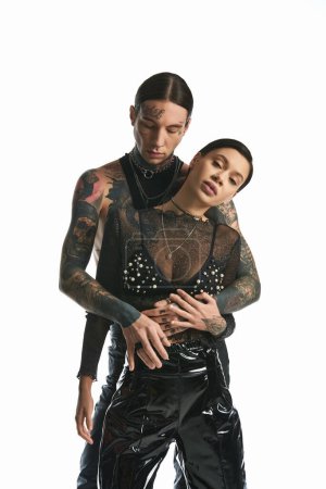 A young man and woman, covered in intricate tattoos, embrace each other in a studio against a grey background.