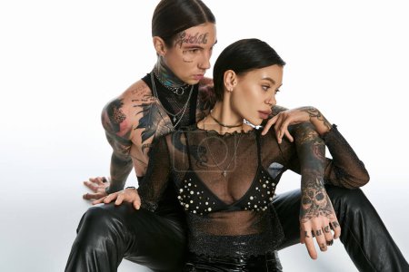 A young tattooed couple sits closely together, sharing a quiet moment of togetherness in a studio against a grey background.