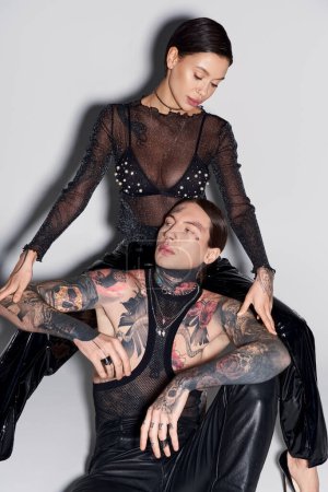 A man with tattoos sitting near woman in a studio setting against a grey background.