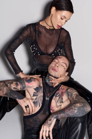 A young woman sits gracefully on the back of a man in a studio setting, both showcasing tattoos, against a grey background.