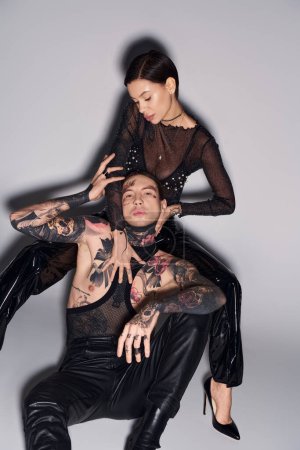 A young woman with tattoos sits gracefully atop a man with tattoos against a grey backdrop in a studio setting.