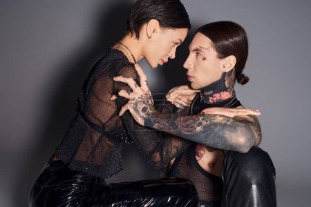 Un jeune couple tatoué assis ensemble dans un studio, partageant un moment de passion et d'intimité sur fond gris.