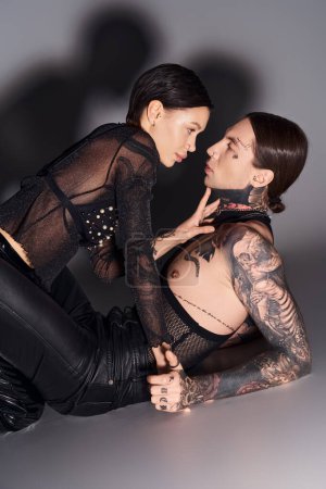A woman with tattoos lays beside a man in a studio against a grey background, connecting in a moment of intimacy.