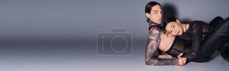 Photo for A young tattooed couple laying side by side on the ground in a studio against a grey background, lost in an intimate embrace. - Royalty Free Image