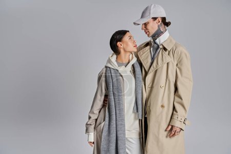 Téléchargez les photos : Un jeune homme et une jeune femme se tiennent côte à côte, tous deux vêtus d'un imperméable, respirant le style et la confiance dans un décor de studio sur fond gris. - en image libre de droit