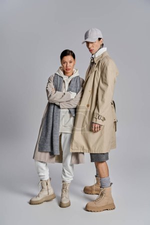 Foto de Un joven y elegante hombre y una mujer parados uno al lado del otro en abrigos de trinchera contra un fondo gris del estudio. - Imagen libre de derechos