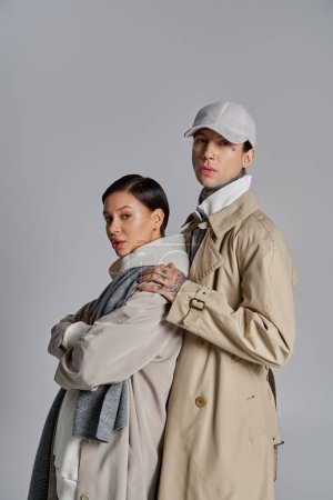 Foto de Un hombre y una mujer jóvenes se paran elegantemente uno al lado del otro en abrigos de trinchera en un estudio sobre un fondo gris. - Imagen libre de derechos