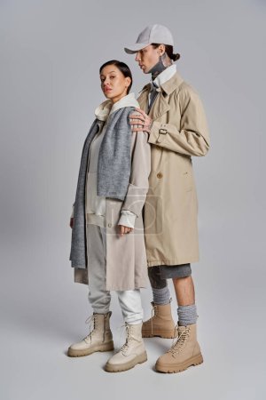 Photo for A young man and woman in stylish trench coats standing closely together in a studio on a grey background. - Royalty Free Image