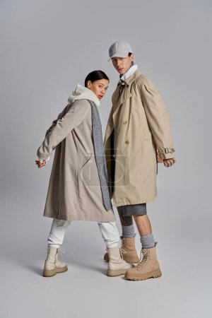 Téléchargez les photos : Un homme et une femme, vêtus de trench coats, se tiennent côte à côte dans un studio au fond gris, respirant élégance et style. - en image libre de droit