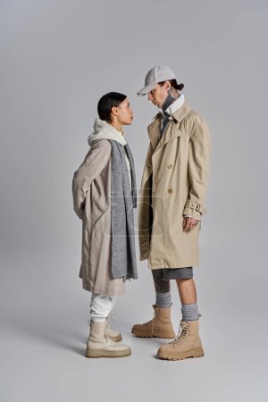 Foto de Pareja de pie lado a lado en abrigos de trinchera con estilo, exudando un ambiente sofisticado y de moda contra un telón de fondo gris. - Imagen libre de derechos