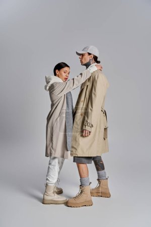 Ein junges stylisches Paar in Trenchcoats steht anmutig zusammen in einem Studio vor grauem Hintergrund.