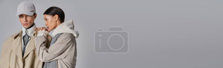 Téléchargez les photos : Un jeune homme et une jeune femme vêtus élégamment de trench coats, debout côte à côte dans un studio sur fond gris. - en image libre de droit