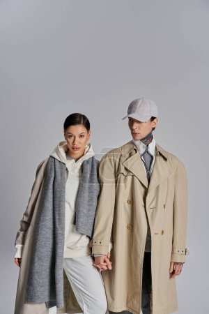 A young stylish couple in trench coats stands gracefully next to each other in a studio set against a grey background.