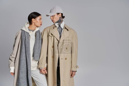 Foto de Un joven hombre y una mujer se paran uno al lado del otro en abrigos de trinchera, exudando elegancia y estilo contra un fondo gris del estudio. - Imagen libre de derechos