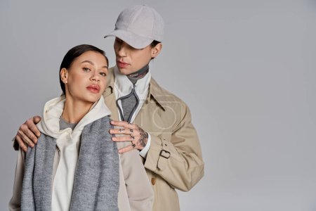 Foto de Un joven hombre y una mujer, ambos vestidos con estilo en abrigos de trinchera, se paran uno al lado del otro en un estudio sobre un fondo gris. - Imagen libre de derechos