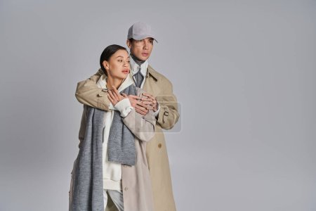 Foto de Un joven y una mujer de moda que usan abrigos de trinchera se paran uno al lado del otro en un estudio con un fondo gris. - Imagen libre de derechos