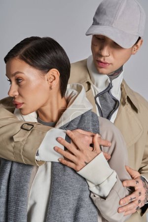 Photo for A young stylish couple in trench coats hold each other tenderly against a grey studio backdrop. - Royalty Free Image