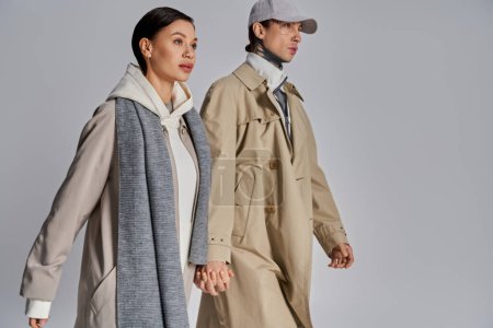 A stylish man and woman confidently walking and wearing trench coats in a studio against a grey background.