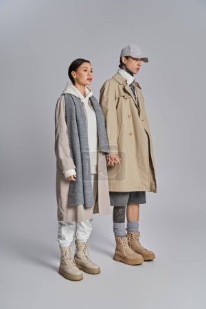 A young stylish couple in trench coats stand next to each other in a studio against a grey background.