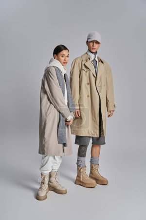 Téléchargez les photos : Jeune couple élégant en trench coats debout côte à côte dans un studio, exsudant l'élégance urbaine sur un fond gris. - en image libre de droit