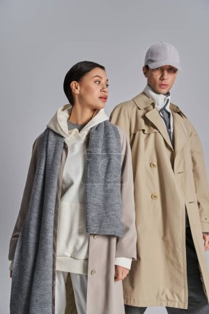 A young stylish couple wearing trench coats and hats in a studio setting against a grey background.
