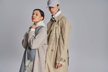 Ein junges stylisches Paar in Trenchcoats steht nebeneinander in einem Studio vor grauem Hintergrund.