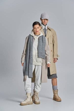 Foto de Un joven hombre y una mujer se paran uno al lado del otro en abrigos de trinchera, exudando estilo y elegancia en un estudio con un fondo gris. - Imagen libre de derechos